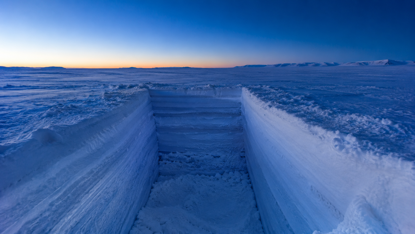 Fosse creusée dans la neige du plateau antarctique, parois blanches stratifiées, sous un ciel bleu de crépuscule.