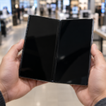 Deux mains tiennent un smartphone pliant ouvert, écran éteint, dans l'allée d'un magasin d'électronique.