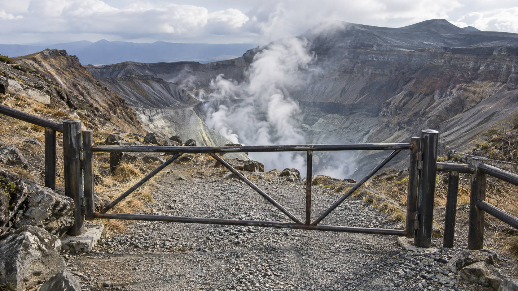 Barrière métallique fermée en travers d’un sentier de gravier au bord d’un cratère volcanique japonais, vapeur au fond.