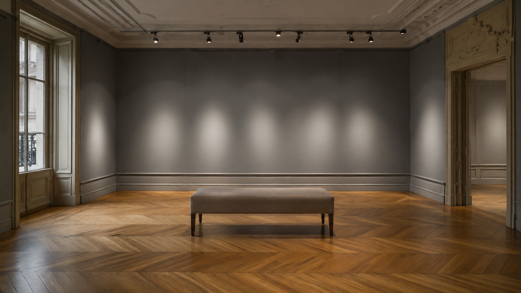 Salle d’exposition vide d’une maison de vente parisienne : cimaise grise éclairée, parquet en point de Hongrie, banquette au centre.