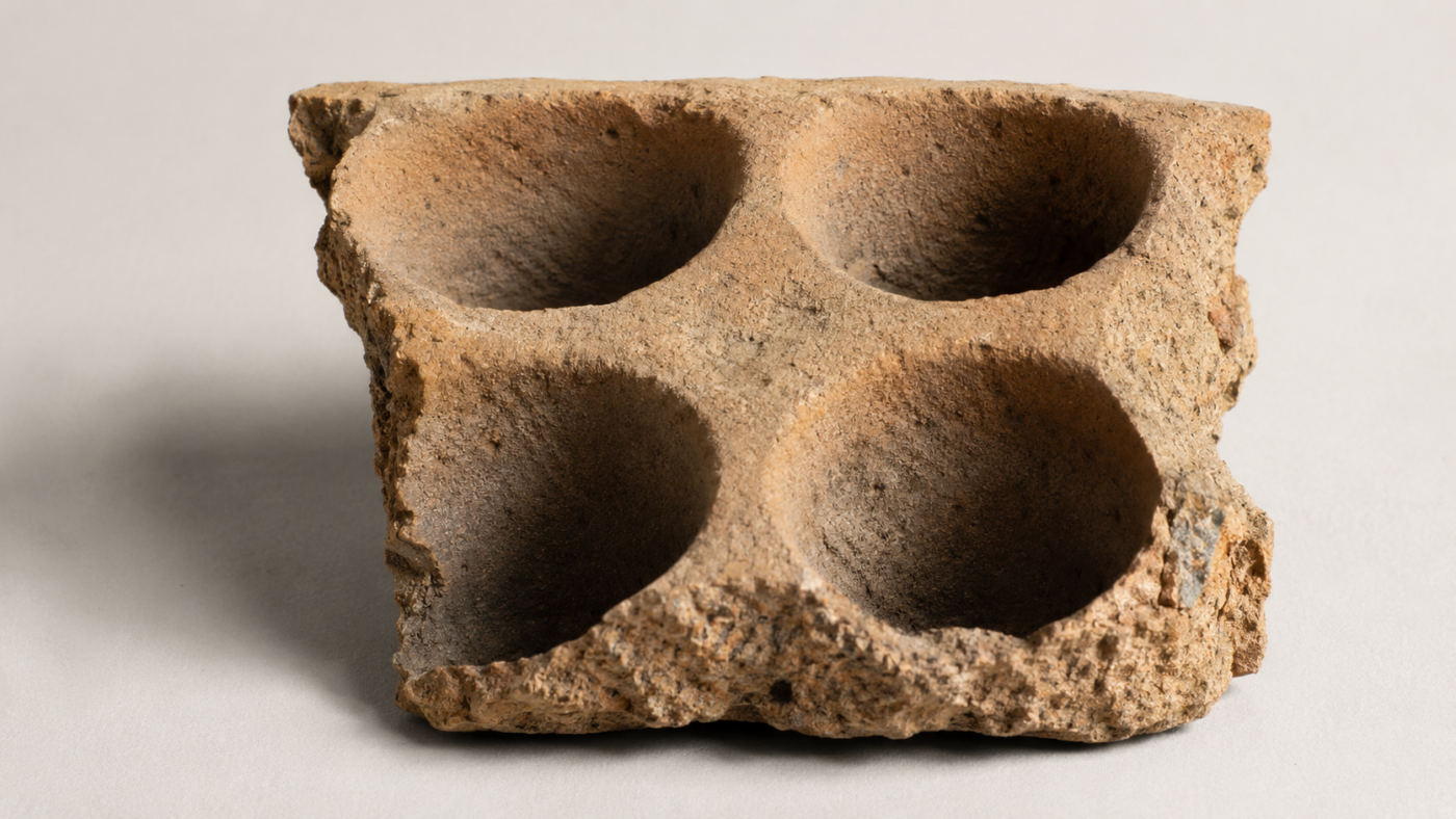 Fragment de plaquette en terre cuite beige percé de quatre alvéoles circulaires à fond évasé, posé sur un fond neutre d’étude.