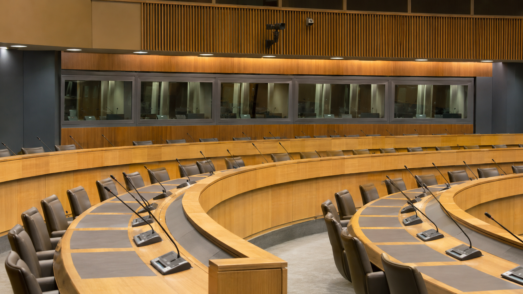 Salle de conférence internationale vide, rangées incurvées de pupitres en bois clair avec micros et cabines d’interprétation vitrées au fond.