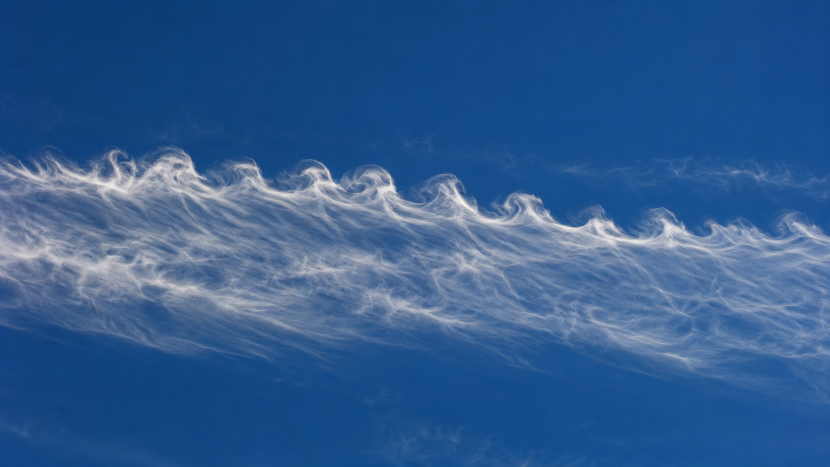 Bande de cirrus fine dans un ciel bleu profond, son bord supérieur enroulé en petites boucles régulières, vue au téléobjectif