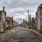 Rue déserte bordée de murs de pierre sans toit dans un village en ruine, sous un ciel couvert.