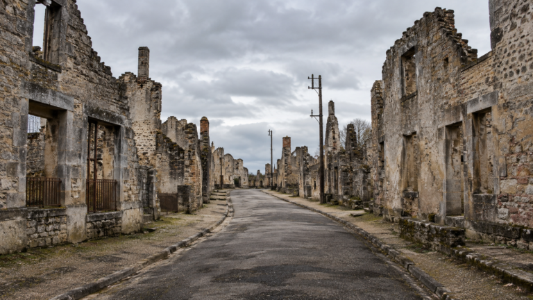 Rue déserte bordée de murs de pierre sans toit dans un village en ruine, sous un ciel couvert.