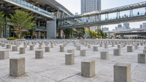 Dalle pavée claire hérissée de petits blocs de béton rectangulaires, devant une gare urbaine japonaise, sous une passerelle piétonne.