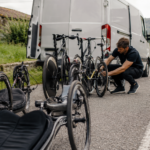 Un mécanicien accroupi aligne des handbikes et des vélos de route adaptés devant un camion d’équipe, sur une route de campagne française.