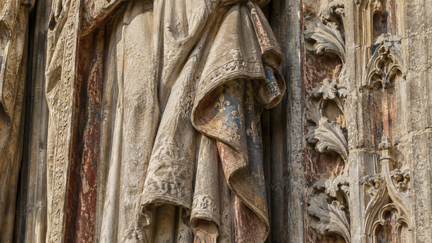 Détail d’un portail gothique en pierre calcaire où subsistent des traces rouges et bleues au creux des plis sculptés.