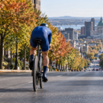 Un coureur cycliste en tenue bleue, vu de dos, en position de contre-la-montre sur une avenue urbaine bordée d'érables.