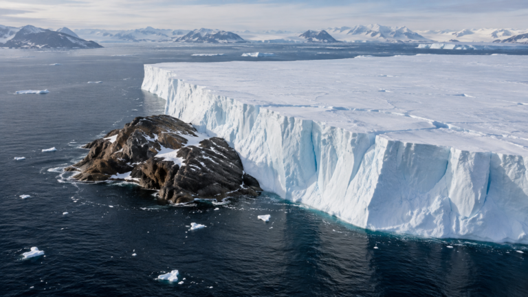 Vue aérienne d’une immense plaque de glace tabulaire calée contre un petit îlot rocheux brun dans un détroit arctique