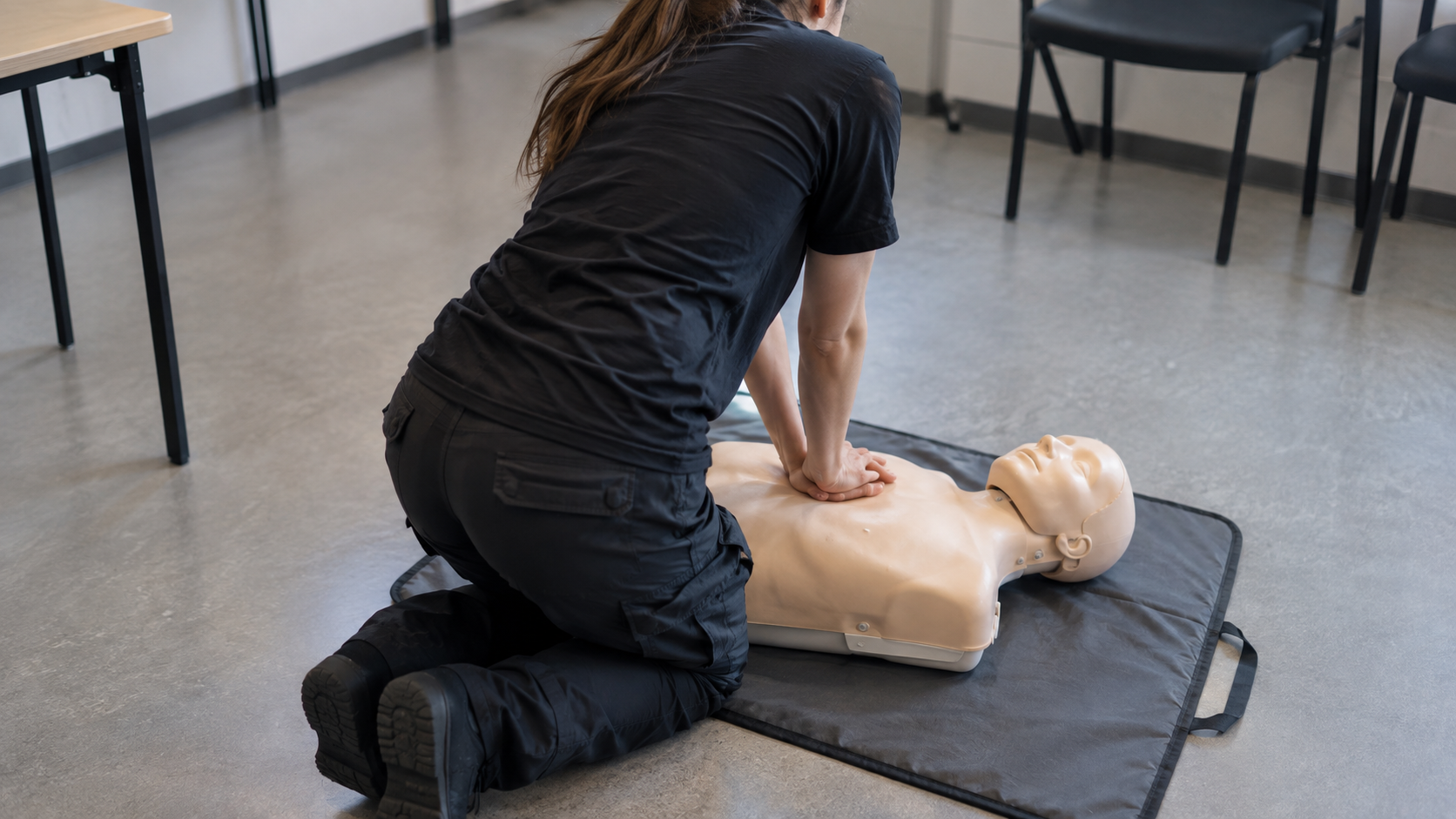 Des mains exercent un massage cardiaque sur un mannequin d’entraînement posé au sol, dans une salle de classe nue.
