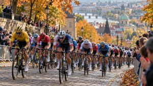 Un peloton de cyclistes grimpe une côte urbaine bordée d’arbres d’automne, derrière des barrières et un public flou.