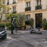Quatre appareils de musculation d’extérieur installés sur le bitume d’une cour d’immeuble parisien, sous de hautes fenêtres.
