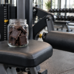 Un bocal ouvert rempli de carrés de chocolat noir, posé sur le siège d'une machine à extension de jambes dans une salle de sport.