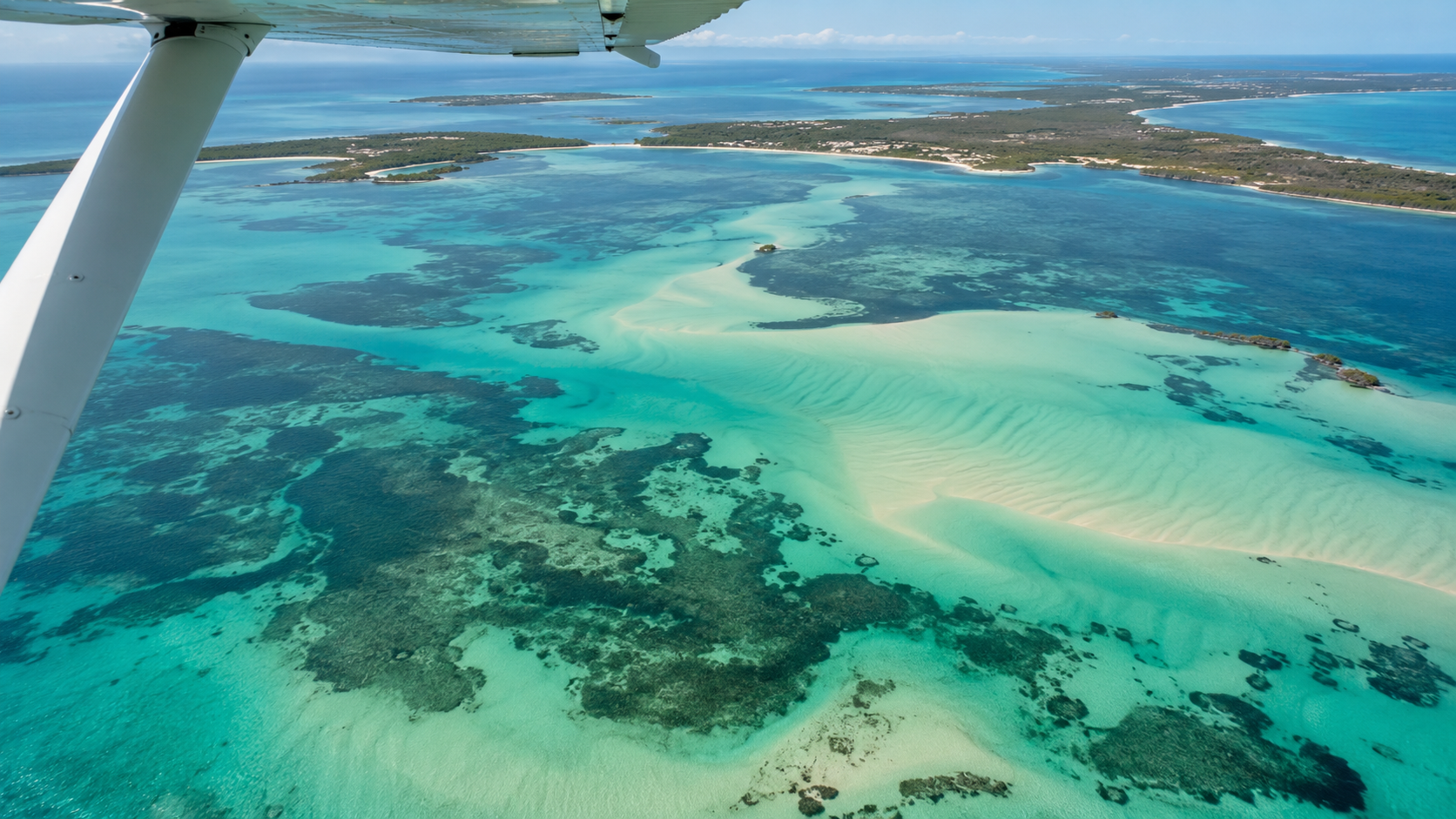 Vue aérienne d'une baie peu profonde turquoise depuis un petit avion, bancs de sable clairs et herbiers sombres sous la surface, hauban d'aile au bord du cadre