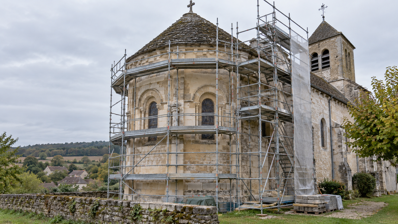Un échafaudage de restauration dressé contre l’abside en pierre d’une petite église rurale française, un matin couvert.