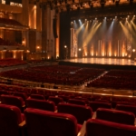 Une grande salle de concert vue des gradins, scène vide éclairée par des projecteurs chauds, sièges rouges au premier plan.