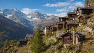 Village de montagne valaisan aux chalets de bois sombre et toits de pierre, accroché à une pente ensoleillée face aux Alpes.