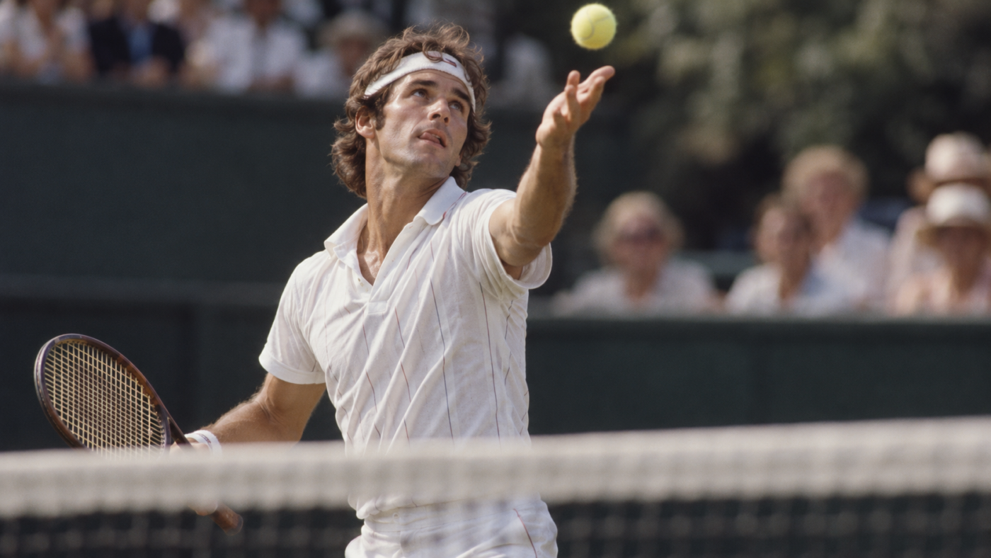 Gros plan sur un joueur de tennis au moment du lancer de balle, bouche entrouverte, vu depuis l’autre côté du filet.