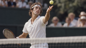 Gros plan sur un joueur de tennis au moment du lancer de balle, bouche entrouverte, vu depuis l’autre côté du filet.