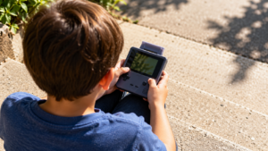 Des mains d’enfant tiennent une console portable en plein soleil, la cartouche dépassant du logement, sur une marche en béton.