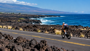 Un triathlète à vélo sur une route qui traverse des champs de lave noire au bord de l'océan