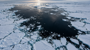 Vue aérienne d’une large étendue d’eau sombre ouverte au milieu de la banquise blanche craquelée, sous une lumière basse d’hiver.