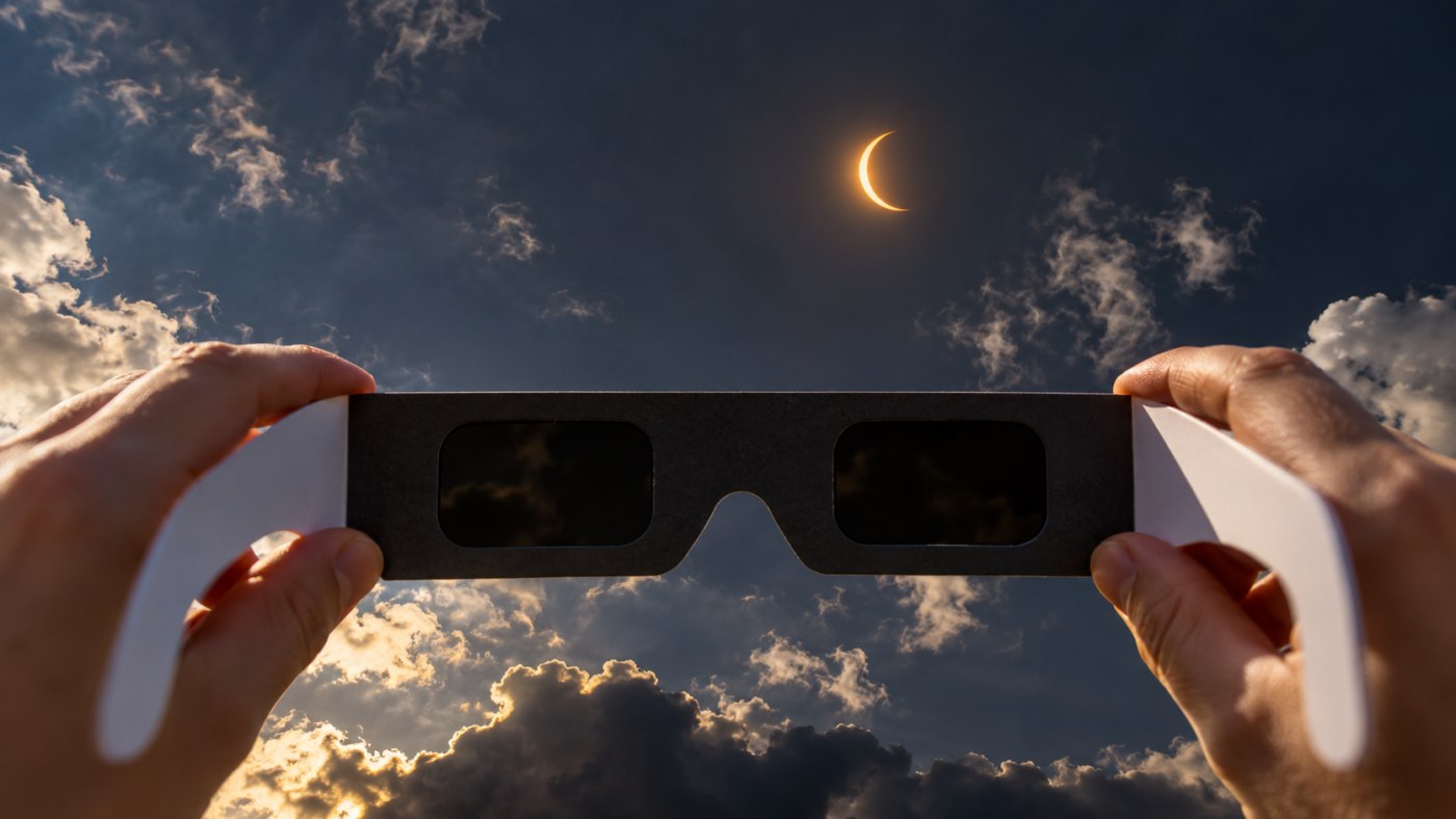 Un mince croissant de Soleil dans un ciel assombri, des lunettes d’éclipse en carton tenues au premier plan